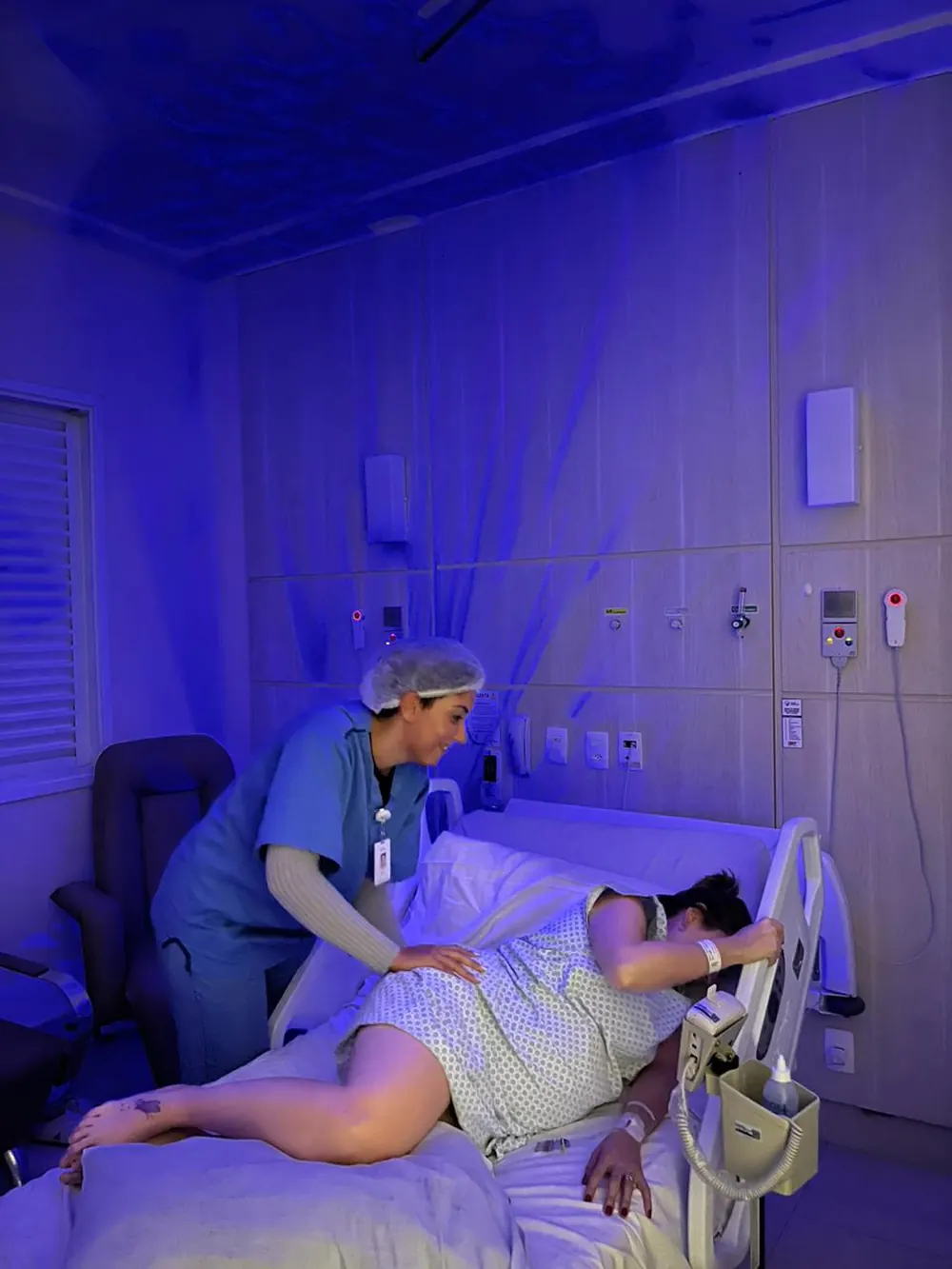 Mulher em trabalho de parto deitada de lado, com a doula ao lado, em sala com luz azul.