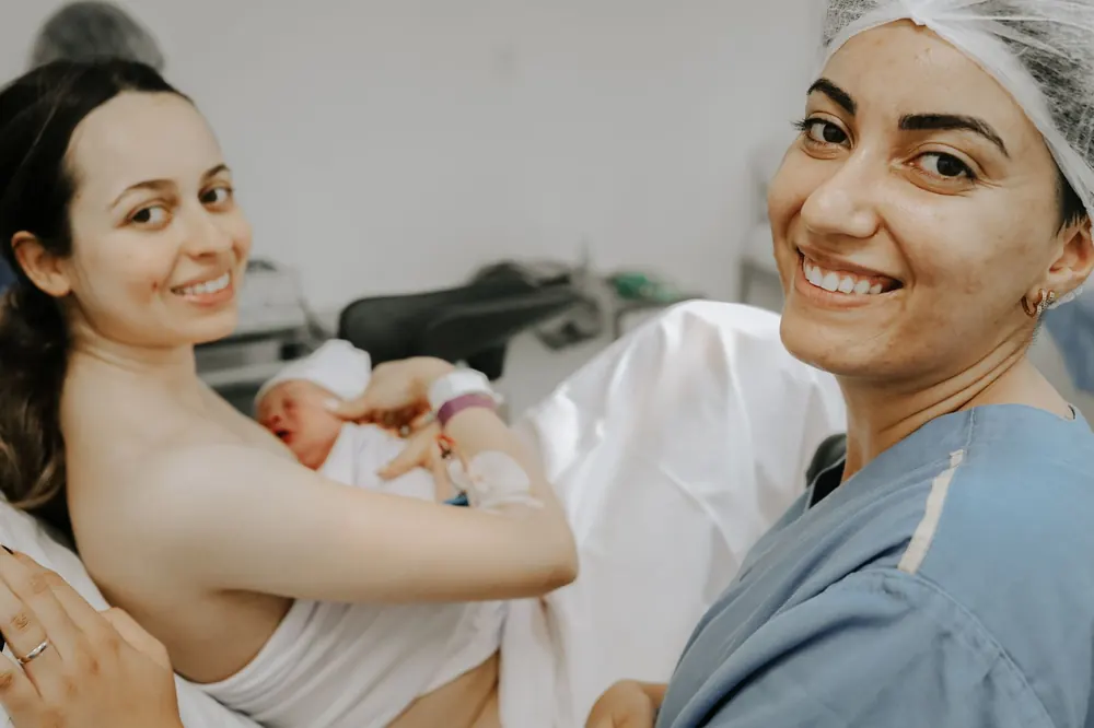 Mãe com o recém-nascido no colo e a doula sorrindo, em selfie na maternidade.